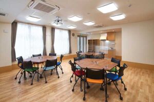 木目調フローリングの広々とした共用スペース。複数のテーブルと椅子が配置され、明るい照明に照らされています。